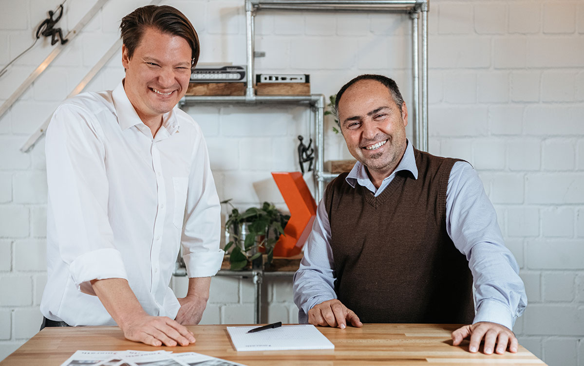 Florian Winger und Giovanni Giordano, die Geschäftsführer von merath metallsysteme, stehen nebeneinander an einem Tisch und lächeln in die Kamera.