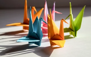 Mehrere farbenfrohe Origami-Kraniche aus Papier stehen auf einer hellen Oberfläche, beleuchtet von warmem Sonnenlicht, das deutliche Schatten wirft