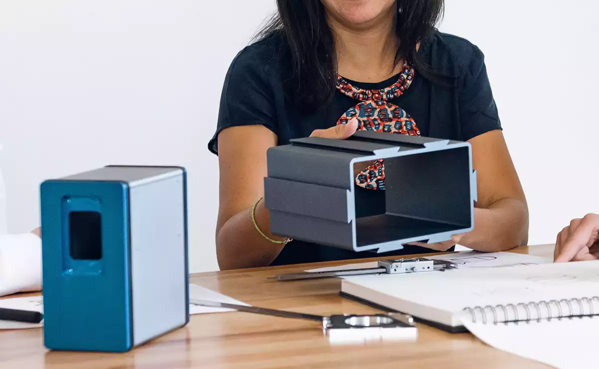 Eine Frau hebt ein Metallgehäuse von einem 3D-Scanner in der Hand. Vor ihr auf dem Tisch steht ein anderes Gehäuse aus Metall mit blauer Frontplatte.