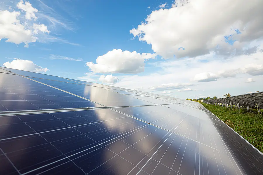 Nahaufnahme schräg aufgestellter Photovoltaikmodule mit Wolkenspiegelung im Glas und grüner Vegetation im Hintergrund.